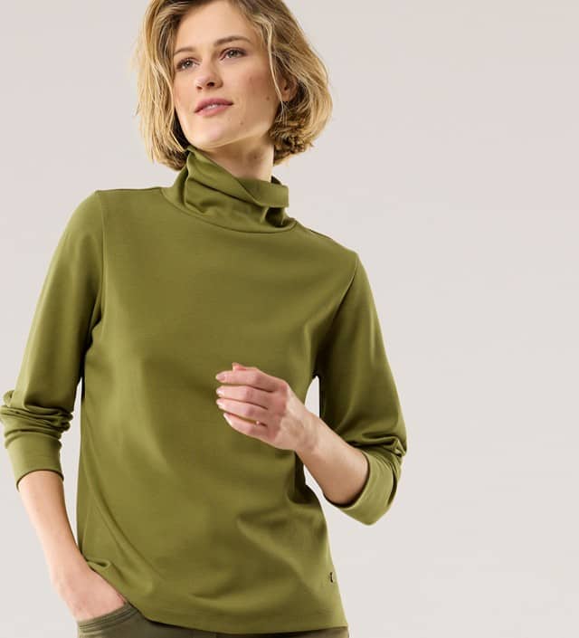 Ein Frau trägt ein Rollkragen-Shirt in Grün und schaut zur Seite.