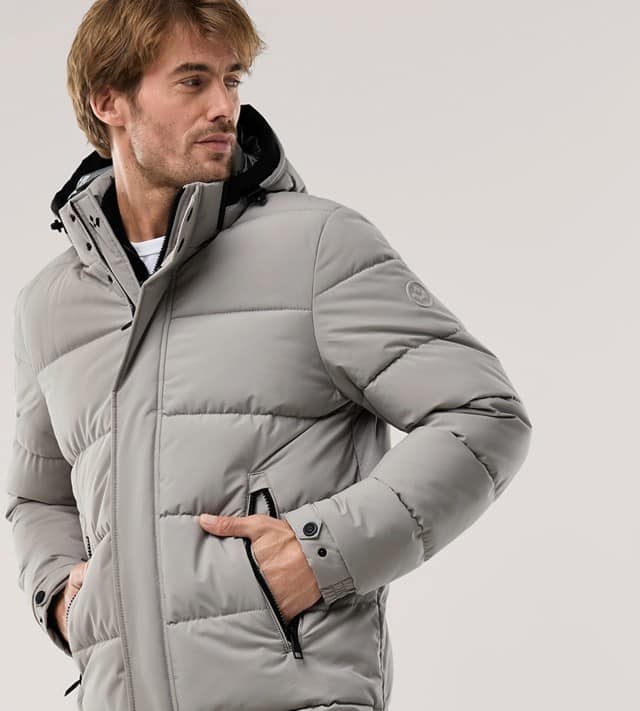 Ein Mann trägt eine Thermojacke in Ecru, hält die Hände in den Taschen und schaut zur Seite.