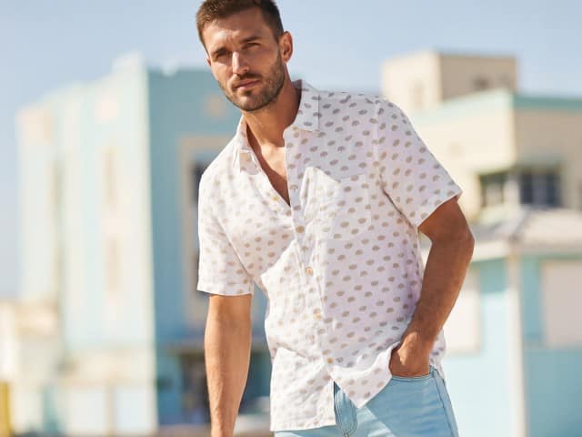 Ein Mann vor einen hellblauen Gebäude, trägt ein sommerliches Kurzarmhemd in weiß mit Muster, dazu hellblaue Shorts.