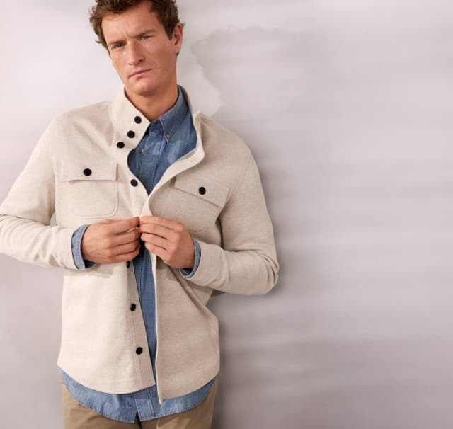 Ein Mann trägt eine naturfarbene Sweatjacke mit Stehkragen, darunter ein blaues Chambray-Hemd.