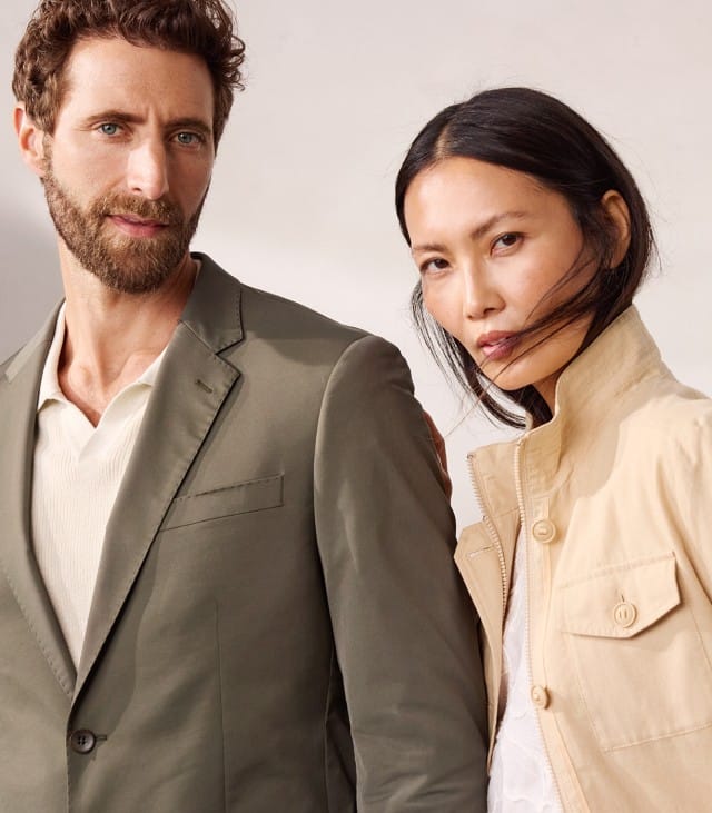 Ein Mann in olivgrünem Anzug und eine Frau in beiger Jacke posieren modern und elegant vor einem hellen Hintergrund. Beide tragen schlichte, stilvolle Kleidung in neutralen Farben.