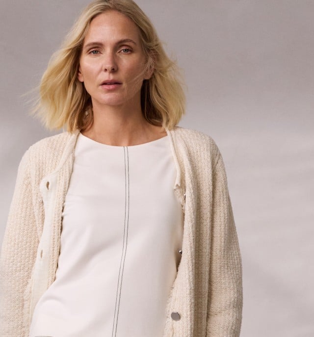 Eine Frau trägt eine Fancy Longstrickjacke in Creme und darunter ein Edeljerseyshirt mit Zierstich in Weiß.
