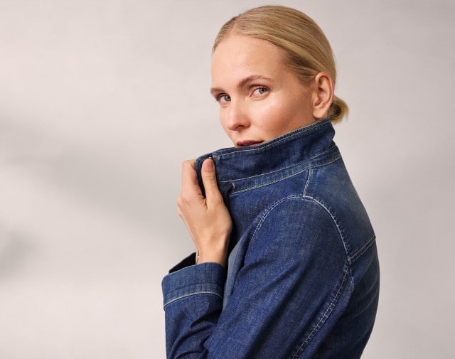 Ein Frau trägt eine hochgeschlossene dunkelblaue Jeansjacke und hält den Kragen fest.