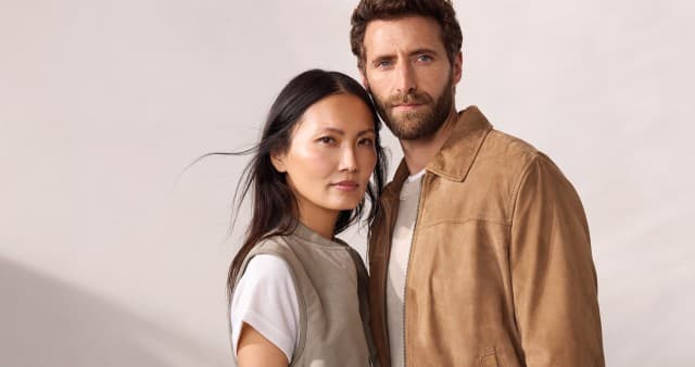 Ein Frau trägt eine Jacquardweste in Khaki, darunter ein weißes Shirt. Ein Mann in einem Ziegenvelours Blouson in Beige und einem Premium Leinen-Pullover in Natur.