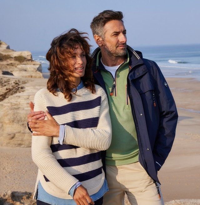 Mann mit blauer Jacke und Frau mit blau-weiß gestreiftem Pullover stehen am Strand