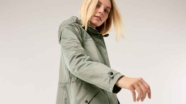 Blonde Frau in grüner Jacke mit Kapuze, blickt nach unten. Hintergrund ist dezent und hell.