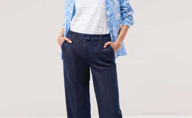 Frau in lässiger Kleidung: weißes T-Shirt, hellblaue Bluse mit Blattmuster, dunkelblaue weite Hose und weiße Sneakers. Hände in Hosentaschen.