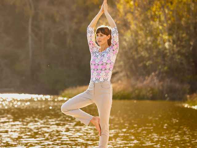 Frau in Yoga-Pose "Der Baum" vor einem See, bunte Bluse und beige Hose tragend.