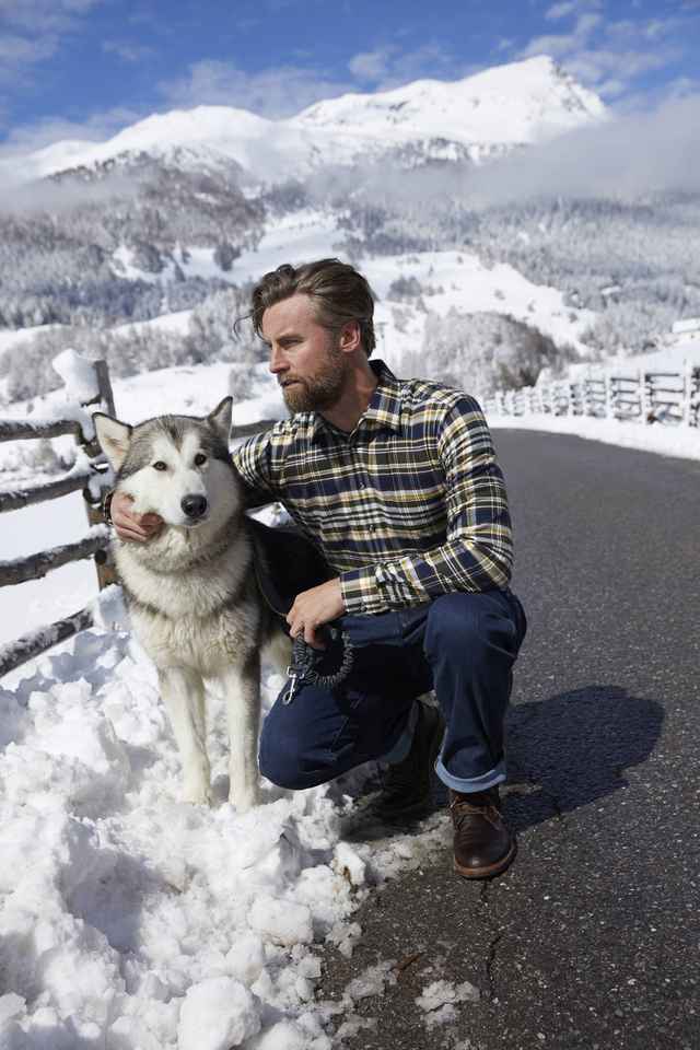 Mann hockt neben einem Husky in einer Schnee Landschaft.