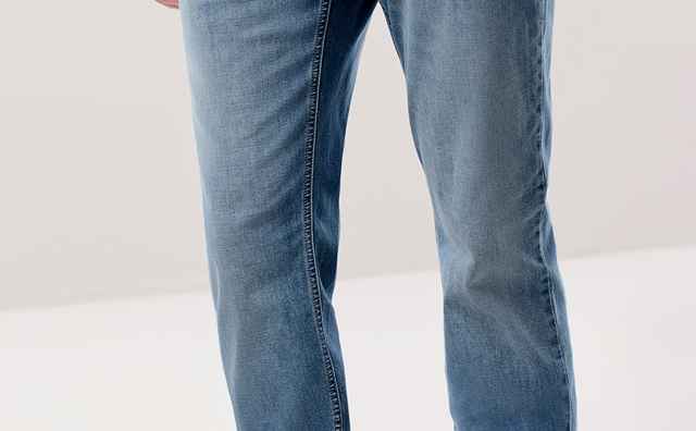 Person trägt eine blaue Jeans und weiße Sneaker, Hände in den Hosentaschen, vor neutralem Hintergrund.