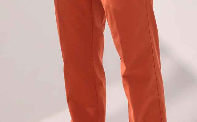 Mann in orangefarbener Hose und weißem T-Shirt, steht locker, Hände in den Taschen, trägt weißen Gürtel und weiße Sneaker.