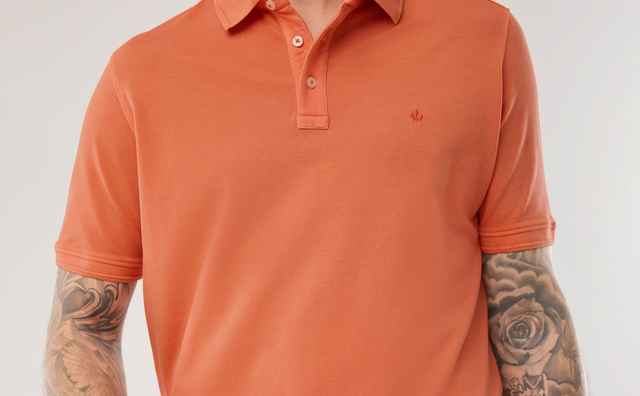 Mann mit orangefarbenem Poloshirt und beigen Hosen posiert, zeigt tätowierte Unterarme, lächelt leicht.