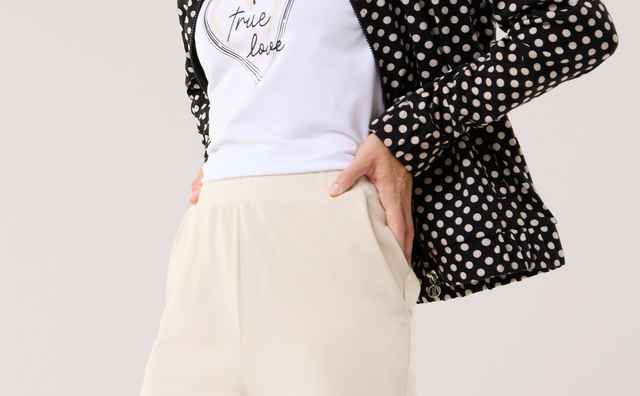 Eine Frau trägt eine schwarz‑weiß gepunktete Jacke über einem weißen T‑Shirt mit Herzmotiv und heller, weiter Hose. Der Look wirkt modern, lässig und leicht elegant.