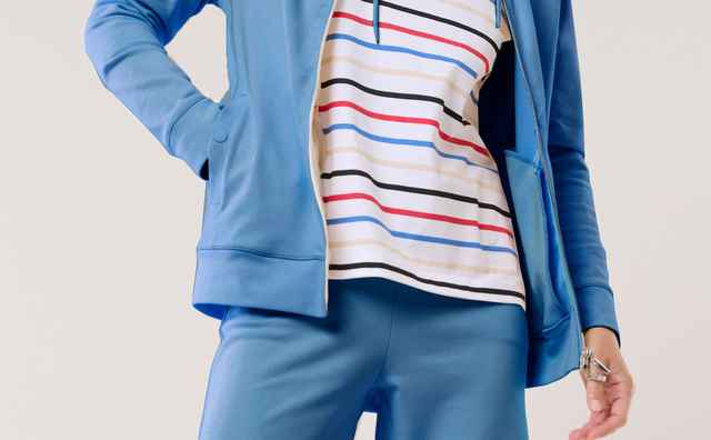 Eine Frau trägt einen blauen Freizeitanzug mit Jacke und weiter Hose, kombiniert mit einem hellen T‑Shirt mit bunten Streifen. Der Look wirkt sportlich und entspannt