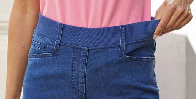 Person zeigt elastischen Bund einer Jeans über einem rosa Shirt.