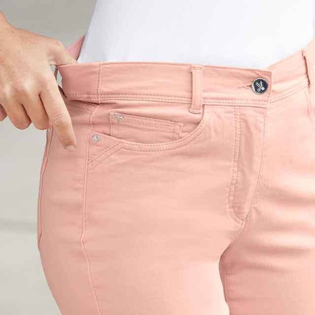 Person trägt eine rosa Hose und ein weißes Oberteil, zeigt mit der Hand auf den Hosenbund.