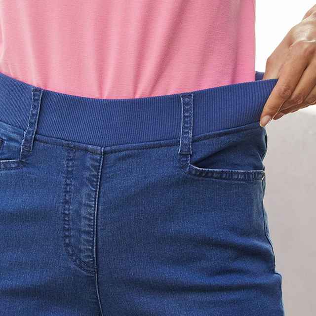 Person trägt eine blaue Jeans und ein rosa Oberteil, zieht an einem elastischen Bund, um die Dehnbarkeit der Hose zu zeigen.