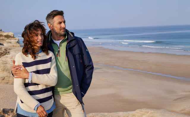 Ein Paar steht am Strand. Sie trägt einen gestreiften Pullover, er eine Jacke. Im Hintergrund das Meer und Klippen.