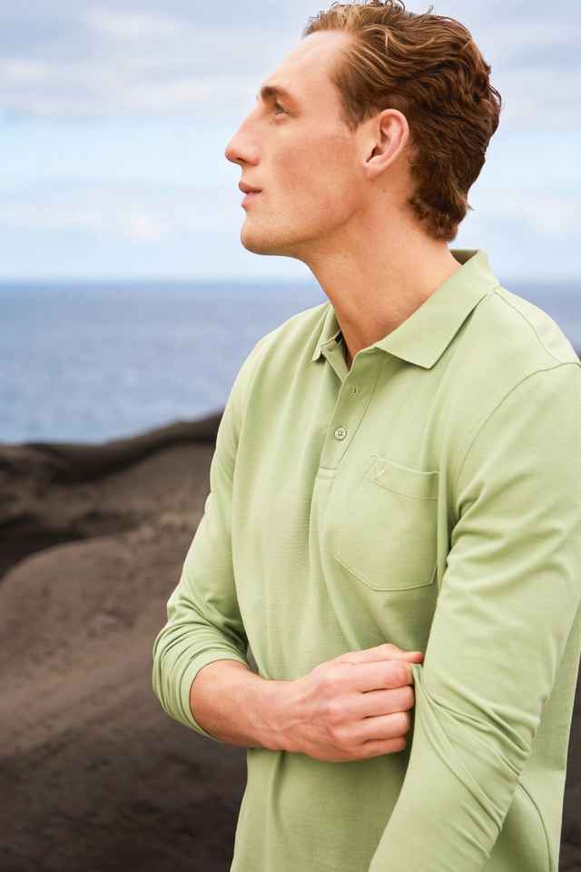 Mann in grünem Polohemd steht im Freien mit Blick auf das Meer, vulkanische Landschaft im Hintergrund, bewölkter Himmel.