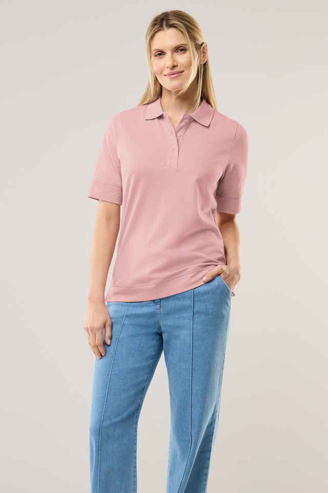 Frau mit blonden Haaren trägt ein rosa Polo-Shirt und blaue Jeans. Sie lächelt und hält eine Hand in der Hosentasche.