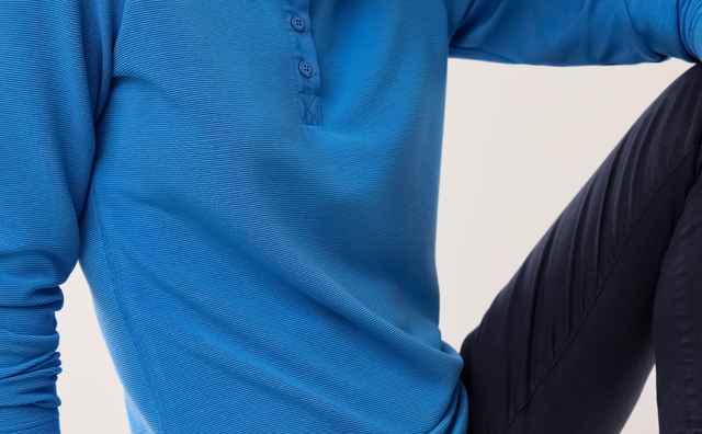 Eine Frau mit blondem Haar trägt ein blaues Poloshirt und dunkle Hosen, sitzt lässig auf einem weißen Block und blickt ernst in die Kamera.