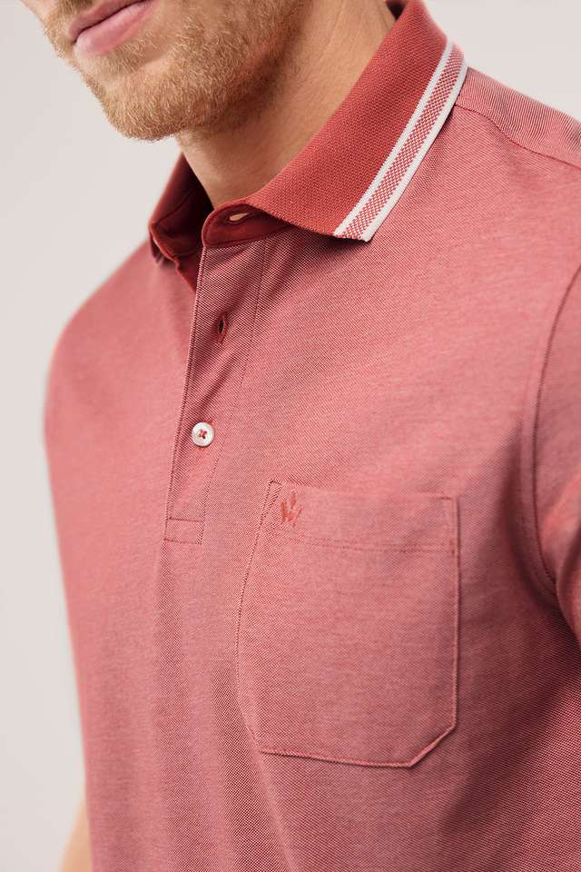 Mann in einem rosa Poloshirt mit Brusttasche und weißen Akzenten, sitzt entspannt und schaut zur Seite vor einem grauen Hintergrund. Angezeigt beim Hover