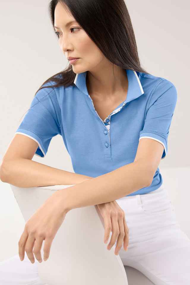 Frau in blauem Poloshirt sitzt auf weißem Stuhl. Sie hat lange Haare und schaut nach unten.