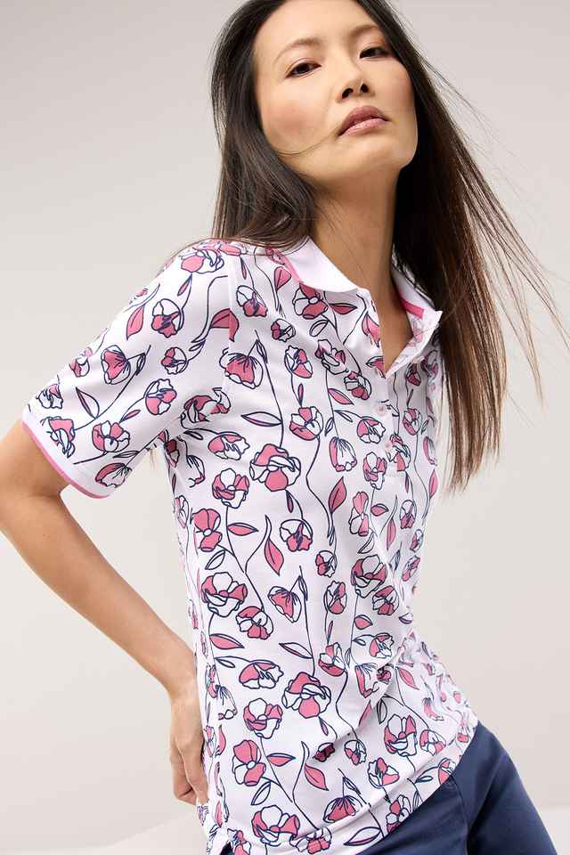 Frau in einem weißen Polo mit rosa Blumenmuster und blauer Hose, lässig posierend vor neutralem Hintergrund.
