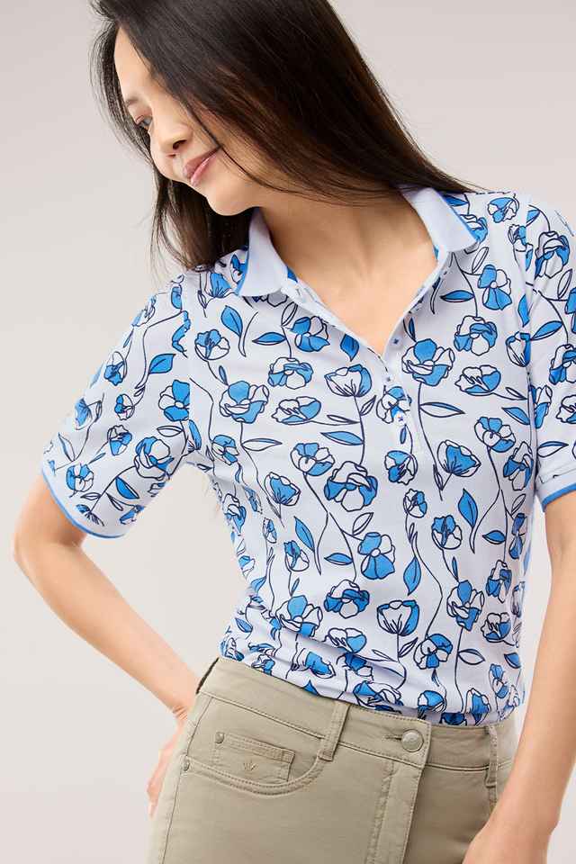 Frau trägt ein hellblaues Poloshirt mit blau-weißem Blumenmuster und eine beige Hose. Sie blickt zur Seite, ihre Haare sind lang und dunkel.