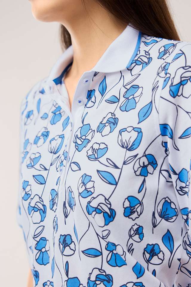 Frau trägt ein hellblaues Poloshirt mit blau-weißem Blumenmuster und eine beige Hose. Sie blickt zur Seite, ihre Haare sind lang und dunkel. Angezeigt beim Hover