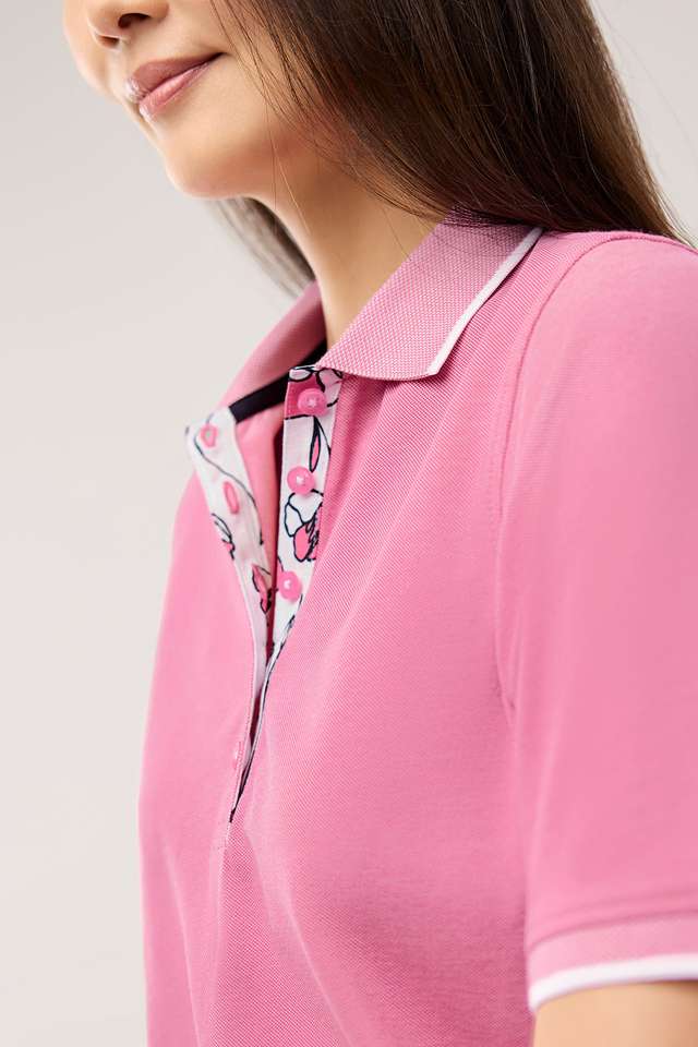 Frau mit langem dunklem Haar trägt ein rosa Poloshirt und weiße Hose. Sie steht vor einem neutralen Hintergrund und blickt in die Kamera. Angezeigt beim Hover