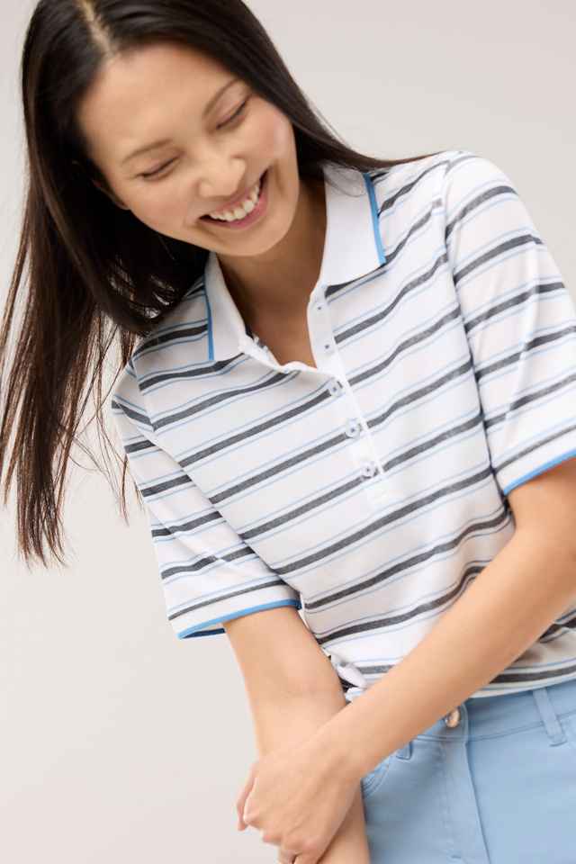 Person mit langen Haaren lächelt und trägt ein gestreiftes Poloshirt mit blauen Akzenten und hellblaue Hose.