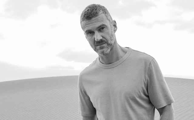 Ein Mann mit Bart steht in einem grauen T-Shirt in einer Wüstenlandschaft, im Hintergrund Sanddünen und bewölkter Himmel. Schwarz-Weiß-Foto.