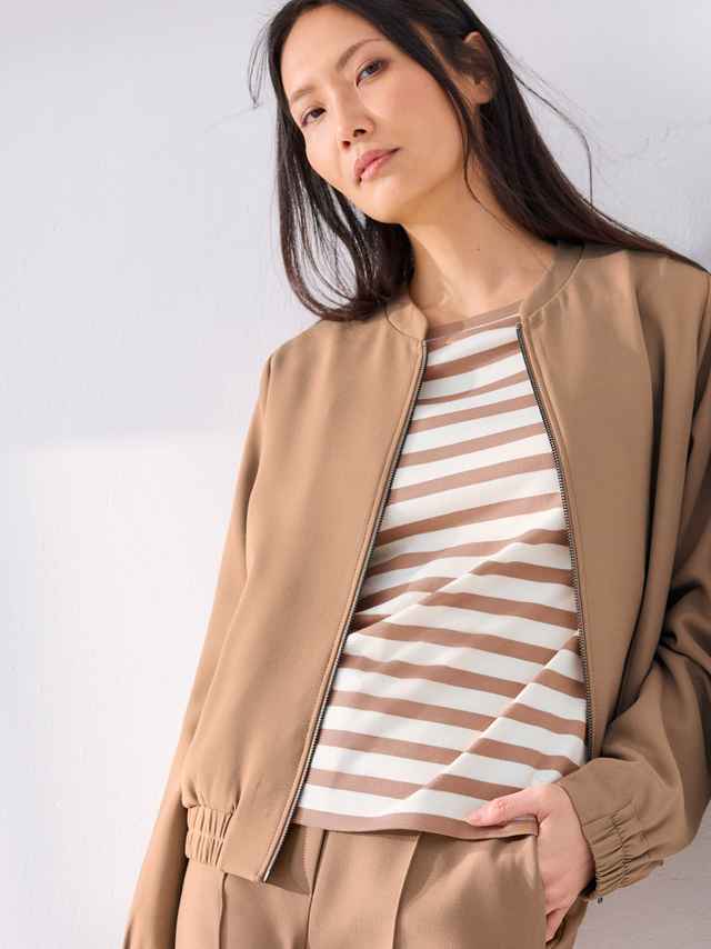 Frau mit langen Haaren in beige Jacke und gestreiftem Shirt posiert gegen eine helle Wand.