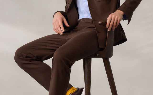 Mann in braunem Anzug und blauer Hemd sitzt auf einem Stuhl, trägt gelbe Socken und braune Slipper, vor grauem Hintergrund.