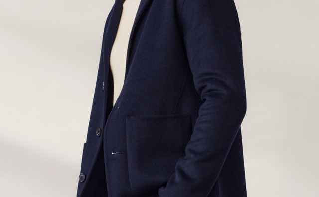 Mann in dunkelblauem Blazer und weißem Rollkragenpullover, steht seitlich mit einer Hand in der Hosentasche vor einem neutralen Hintergrund.