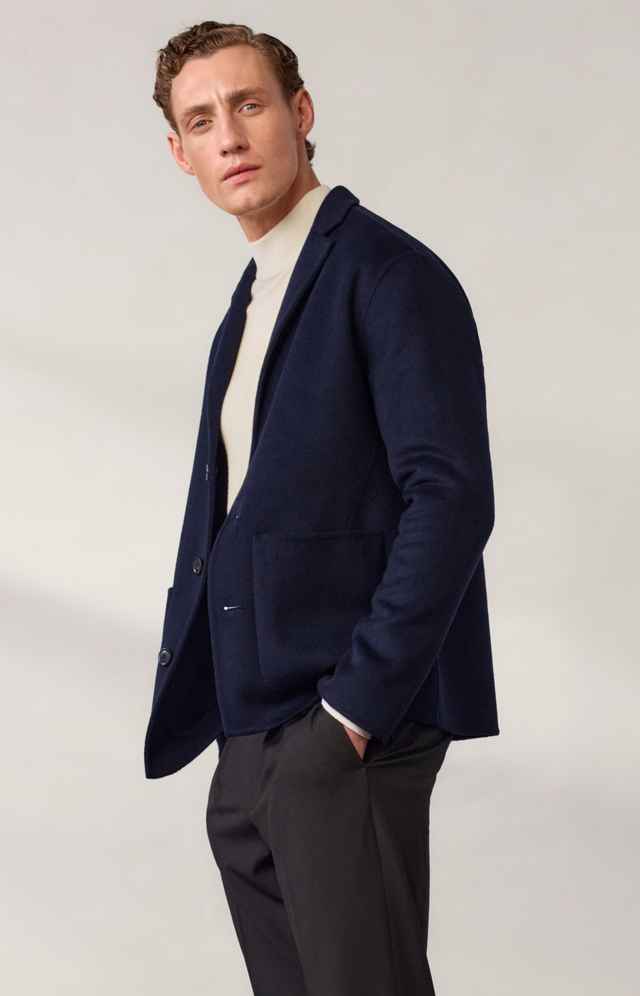 Mann in dunkelblauem Sakko und schwarzer Hose, beige Rollkragenpullover, posierend vor neutralem Hintergrund.