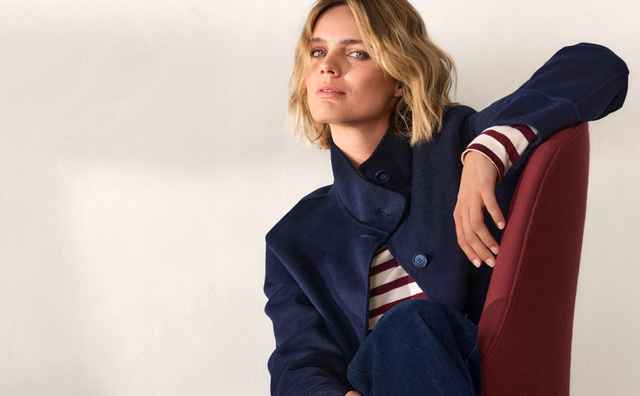 Frau in blauer Jacke und Jeans sitzt lässig auf einem roten Stuhl. Sie trägt ein gestreiftes Shirt und rote Schuhe.