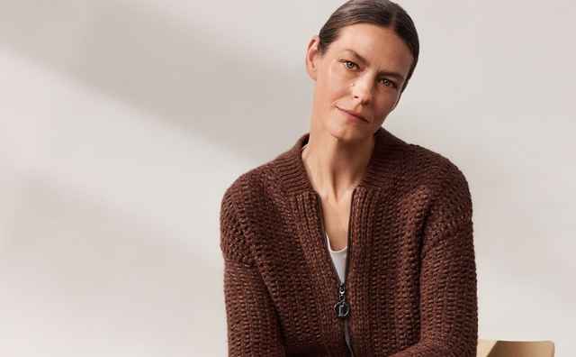 Person in brauner Strickjacke und grauer Hose sitzt auf einem Stuhl, Beine übereinandergeschlagen, in einem minimalistischen Raum.