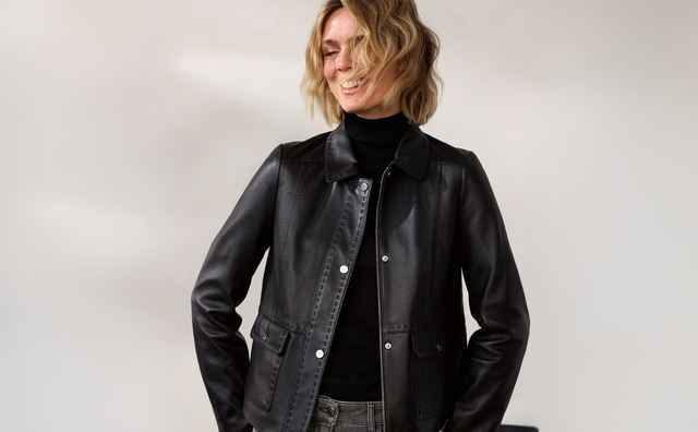Frau mit blonden Haaren lächelt in die Kamera. Sie trägt eine schwarze Lederjacke, ein schwarzes Oberteil und graue Hose. Hintergrund: minimalistisch.