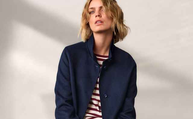 Person mit blondem Haar trägt ein gestreiftes Shirt und eine dunkelblaue Jacke, sitzt vor einer hellen Wand.