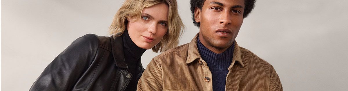 Zwei Personen posieren in stilvoller Kleidung; eine trägt eine schwarze Lederjacke, die andere ein braunes Cordhemd. Neutrale, helle Kulisse.