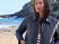 Frau in einer gesteppte Jacke steht am Strand mit windgepeitschten Haaren, im Hintergrund sind Felsen und das Meer zu sehen.