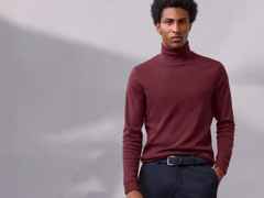 Ein Mann trägt einen bordeauxroten Rollkragenpullover und dunkelblaue Hose, vor einem grauen Hintergrund. Er schaut direkt in die Kamera.