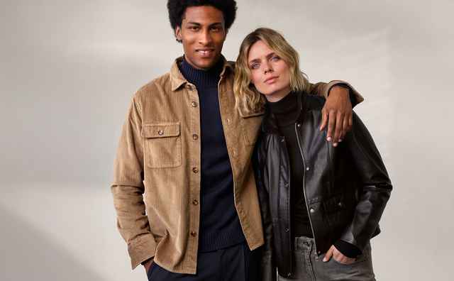 Ein Mann und eine Frau posieren zusammen, er trägt eine beige Jacke, sie eine schwarze Lederjacke; beide schauen in die Kamera.
