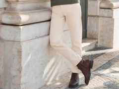 Person in beigefarbenen Hosen lehnt an einer Steinsäule; braune Stiefel, grüner Pullover; entspanntes Ambiente.