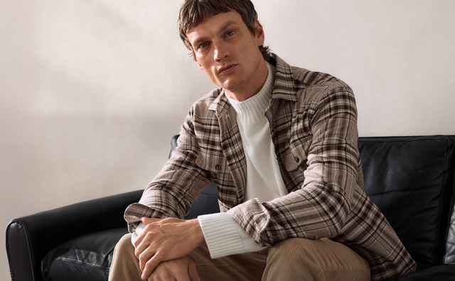 Ein Mann sitzt auf einem schwarzen Ledersofa. Er trägt eine karierte Jacke und einen weißen Pullover. Der Hintergrund ist schlicht und beige.