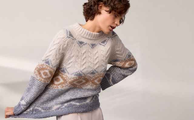 Person mit lockigem Haar trägt einen gestrickten Pullover mit geometrischem Muster in Creme, Blau und Braun vor einem neutralen Hintergrund.