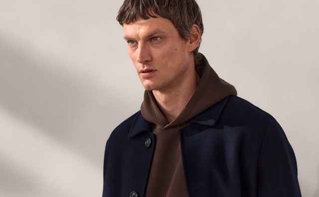 Ein Mann trägt einen dunkelblauen Mantel über einem braunen Hoodie und brauner Hose, steht vor einem grauen Hintergrund und blickt zur Seite.