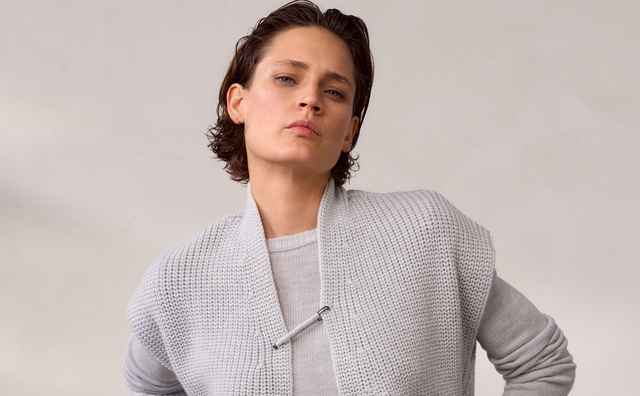 Frau mit kurzem dunklem Haar trägt einen grauen Strickpullover über einem hellen Shirt und blaue Jeans mit Knöpfen, vor neutralem Hintergrund.